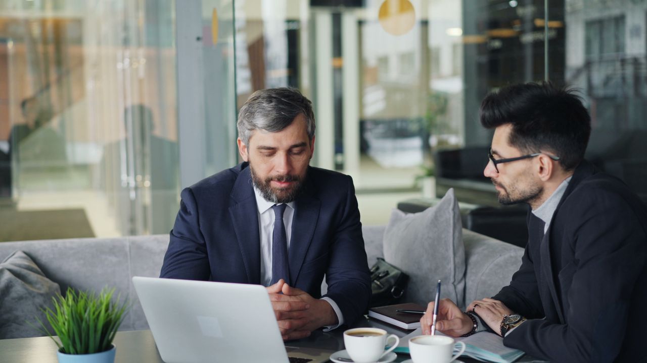 Services Two businessmen in suits discussing work at a cafe with laptops and coffee.
